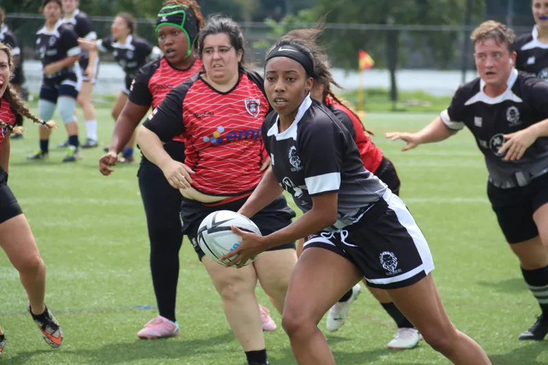A Women's+ Lions ball carrier looks for a pass with a defender in red closing in