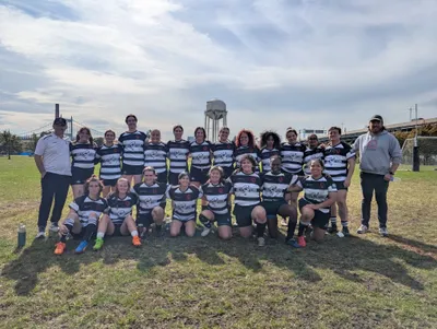 The Women's+ 15s squad in hooped jerseys lined up on the pitch, a water tower and bridge behind them